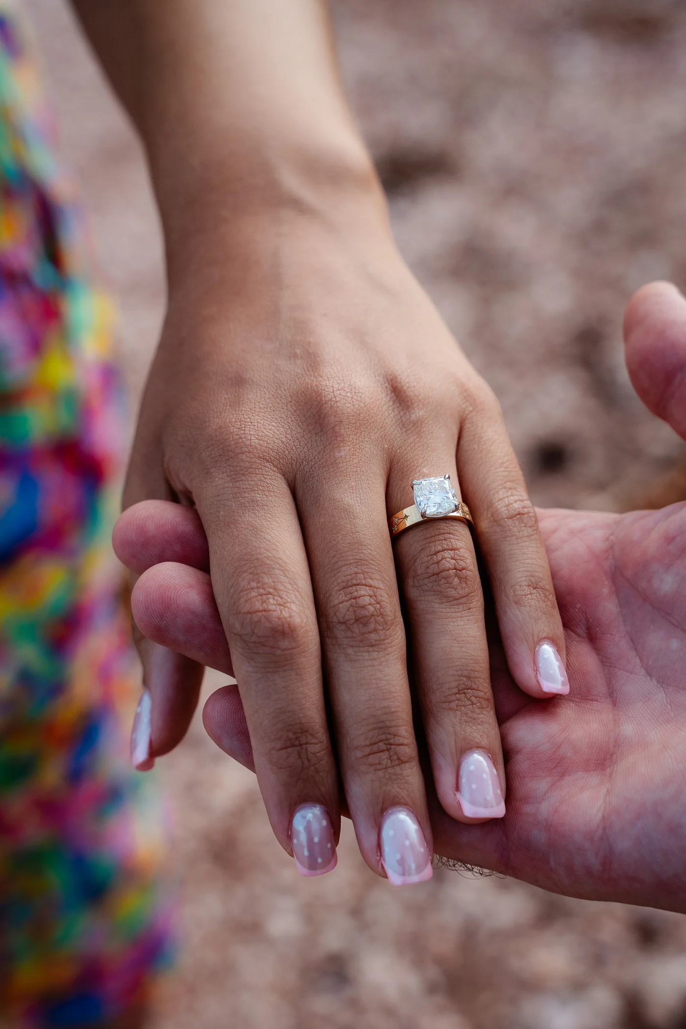 Her hand resting in his palm on red rock in Sardinia, wearing a cushion cut diamond engagement ring on a yellow gold band hand engraved with stars, designed at Haejin Jewelry in Williamsburg, Brooklyn