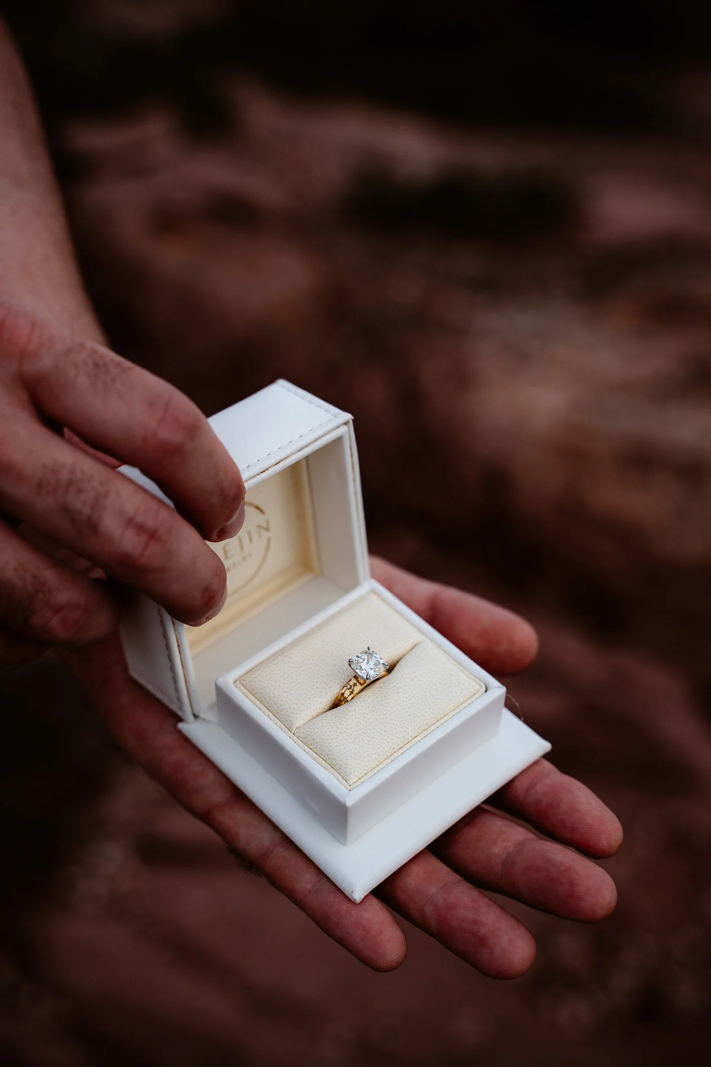Hand holding an open white Haejin Jewelry ring box with a cushion cut yellow gold engagement ring inside, on red rock in Sardinia