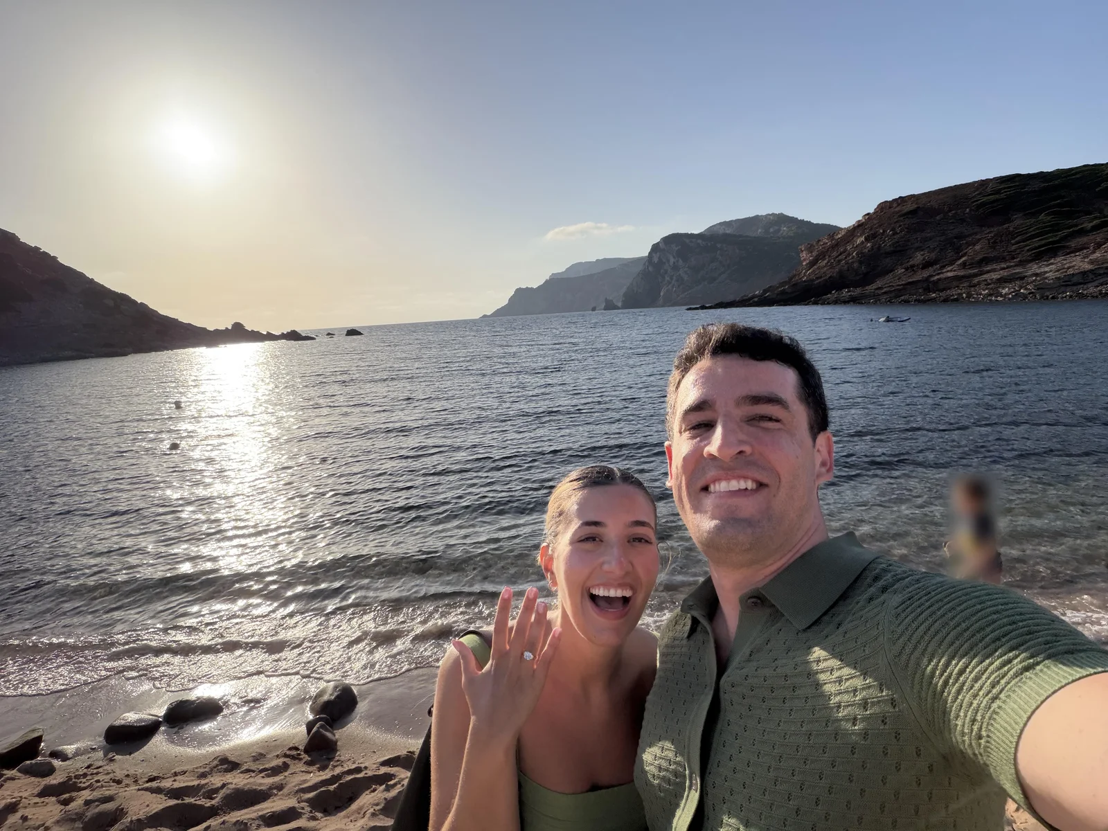 Newly engaged couple laughing on a beach in Alghero, Sardinia at sunset, she is holding up her hand to show a custom cushion cut constellation engagement ring designed at Haejin Jewelry in Williamsburg, Brooklyn
