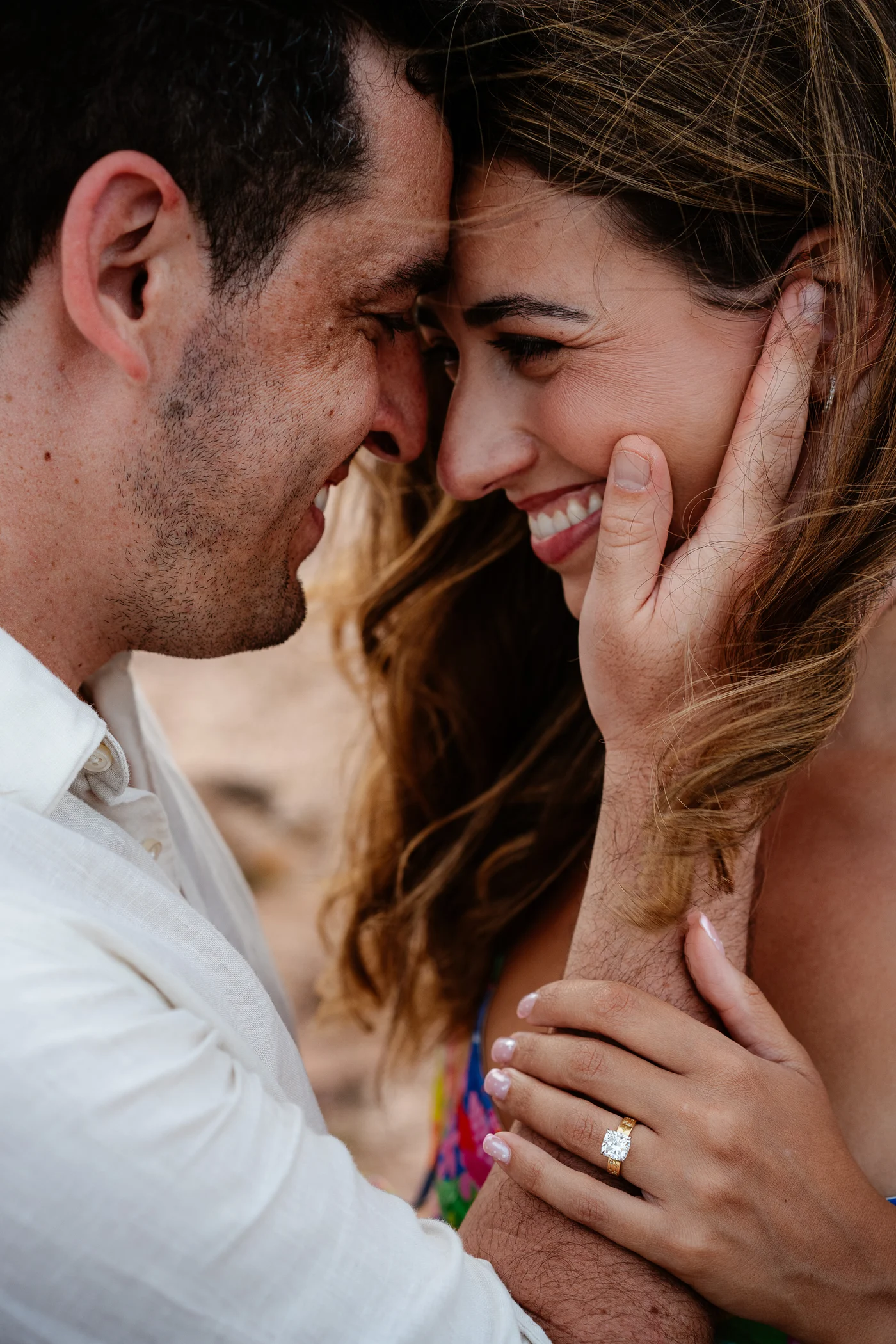 Newly engaged couple laughing forehead to forehead in Sardinia, her hand on his cheek wearing a cushion cut yellow gold engagement ring from Haejin Jewelry
