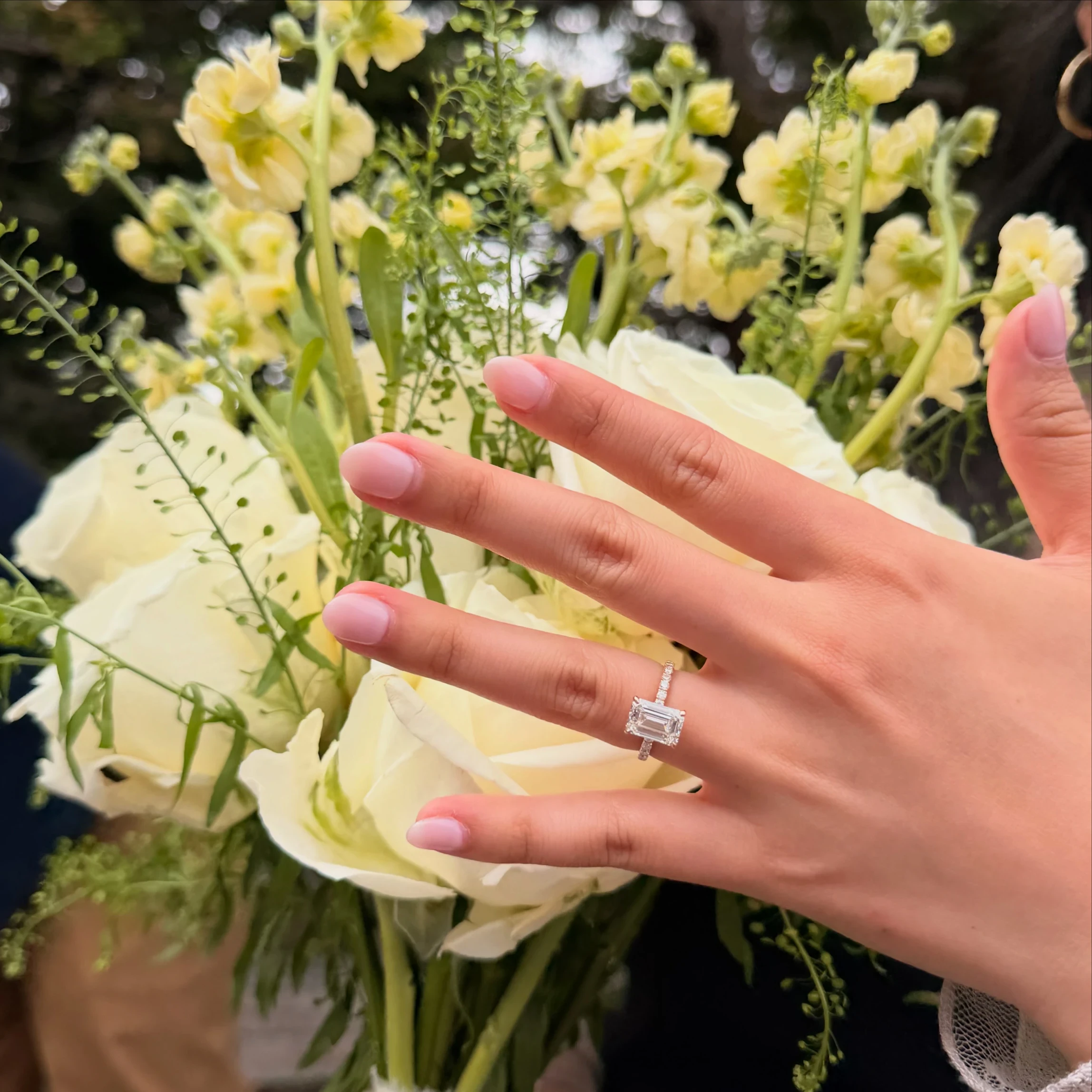 Emerald cut lab-grown diamond engagement ring with pave band on hand with white rose bouquet, Haejin Jewelry customer